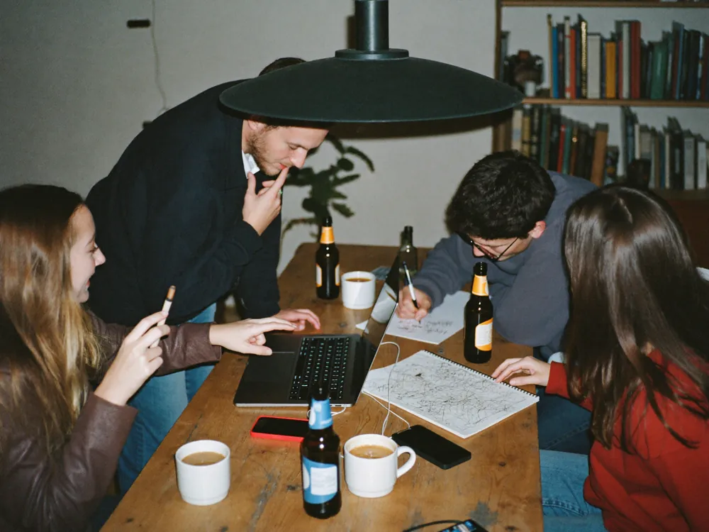 Vier Freunde sitzen abends mit Laptop, Handys und Notizblock um einen Küchentisch und planen